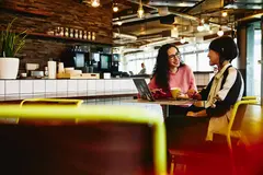 Two female sitting in restaurant having a converstation, looking at laptop, smiling.
