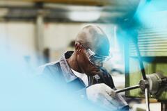 Factory worker operating a column drill. African-American man wearing safety goggles. Primary color blue.